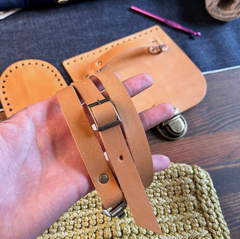 May include: A close-up of a brown leather strap with a brass buckle. The strap is folded in half and held in a hand. The strap is likely for a bag or purse.