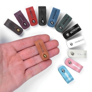 Op de afbeelding: Assortiment van handgemaakte leren kabelbinders in verschillende kleuren, waaronder bruin, rood, blauw, groen, roze en wit. Elke kabelbinder heeft een metalen ring en het woord "HANDMADE" is erop gegraveerd.