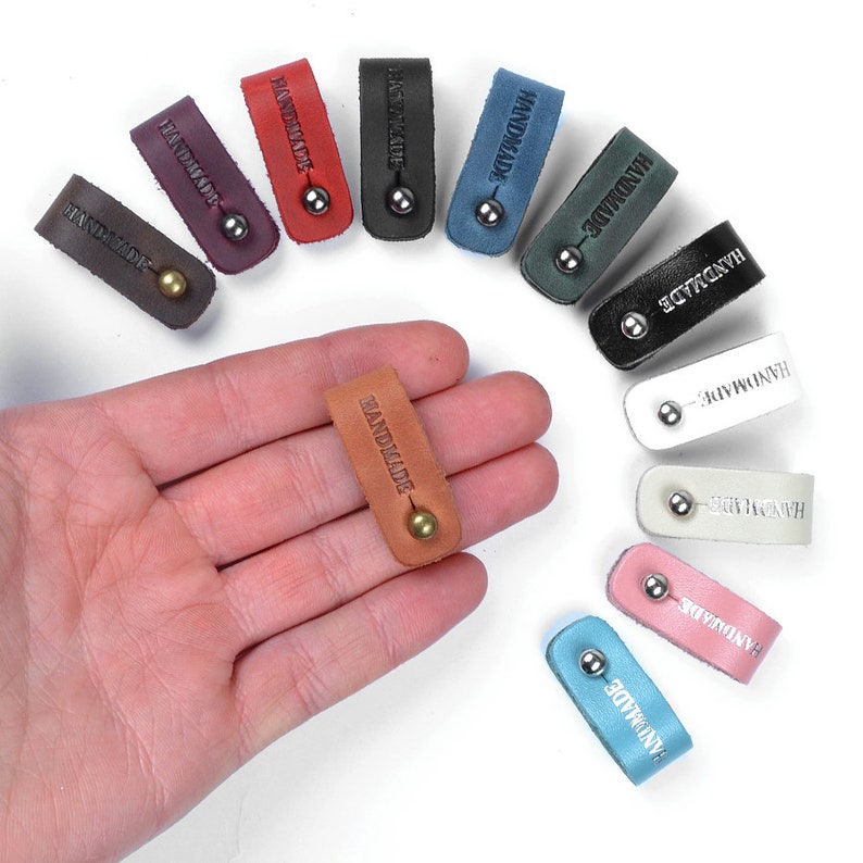 Op de afbeelding: Assortiment van handgemaakte leren kabelbinders in verschillende kleuren, waaronder bruin, rood, blauw, groen, roze en wit. Elke kabelbinder heeft een metalen ring en het woord "HANDMADE" is erop gegraveerd.