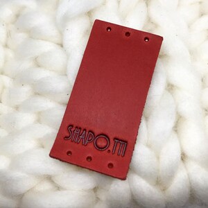 Puede incluir: Etiqueta de cuero roja con el texto "SHAPO.TI" en color morado oscuro.