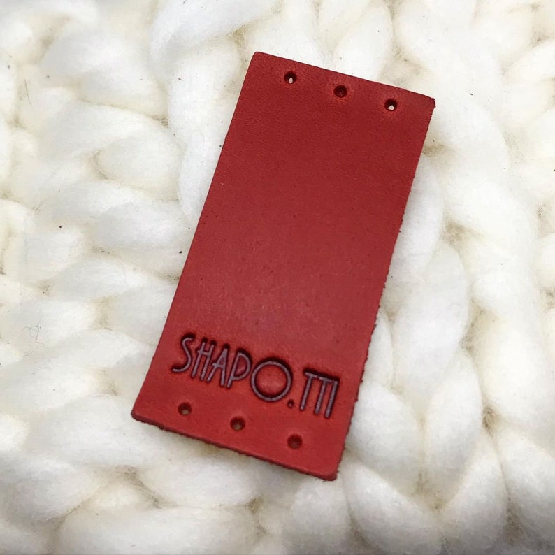 Puede incluir: Etiqueta de cuero roja con el texto "SHAPO.TI" en color morado oscuro.