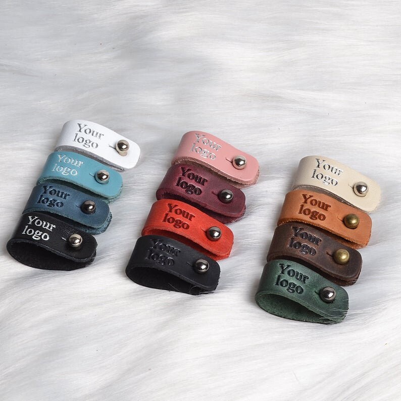 Puede incluir: Surtido de diez organizadores de cables de cuero en varios colores, incluyendo blanco, rosa, rojo, marr&oacute;n, negro y verde. Cada organizador tiene un anillo de metal y el texto "Your logo" grabado en el cuero.