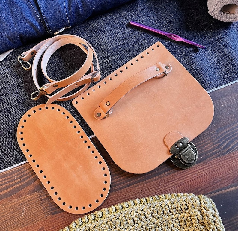 May include: A set of leather crafting supplies including a brown leather bag with a handle and a metal clasp, a brown leather oval shape with holes around the edge, and a brown leather strap with metal hardware.