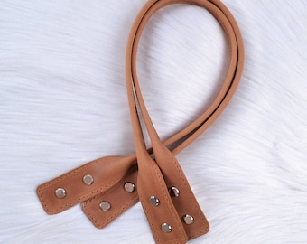 Puede incluir: Dos correas de cuero marrón con remaches de metal plateado. Las correas están conectadas en la parte superior y tienen un bucle en la parte inferior.