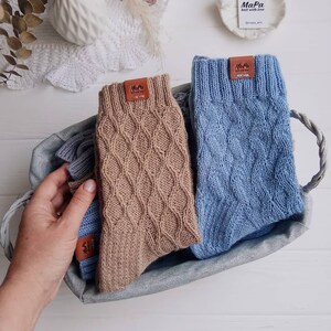 Puede incluir: Dos pares de calcetines de punto en una cesta tejida. Un par es marr&oacute;n y el otro es azul. Ambos pares tienen una etiqueta de cuero con el texto "MaPa knit with love @mapa_knit".