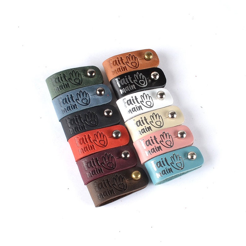 May include: A collection of 12 leather cord keepers in various colors, each with the text "Fait Main" and a hand with a heart embossed on them.