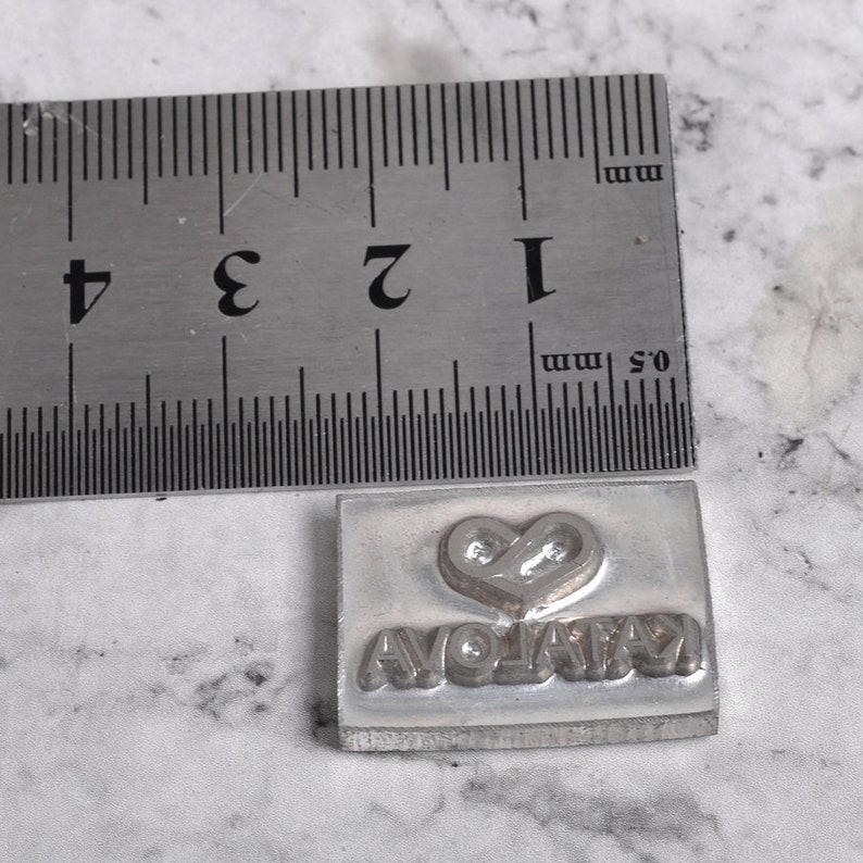 May include: A silver metal stamp with a heart and the word "AVOLATA" engraved on it. The stamp is about 1 inch wide and 1 inch tall.