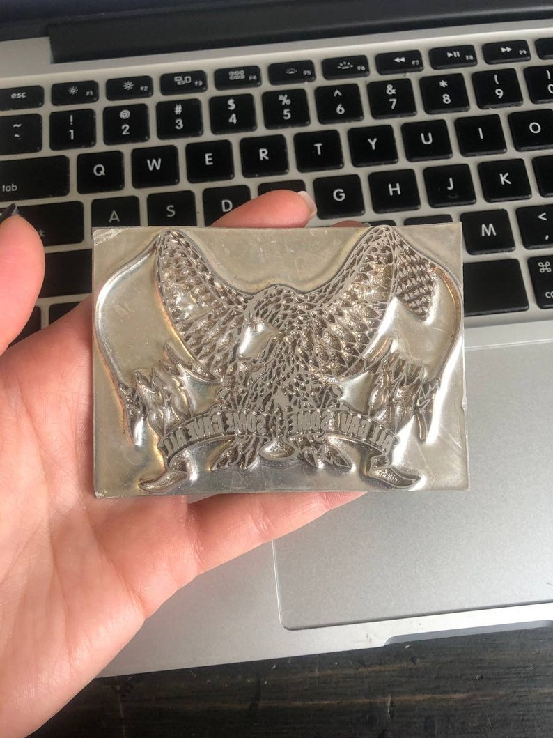 May include: A silver metal stamp with an eagle design and the text "Give Some Wings" around the eagle.