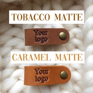 Puede incluir: Dos etiquetas de cuero con el texto "Your logo" en relieve. La etiqueta superior es de color tabaco mate y la etiqueta inferior es de color caramelo mate.
