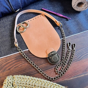 May include: A light brown leather pouch with a brass clasp and chain. The pouch is attached to a long chain with two smaller chains attached to the main chain. The pouch is sitting on a wooden surface.