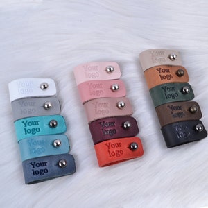 Puede incluir: Surtido de organizadores de cables de cuero en varios colores, incluyendo rojo, naranja, marr&oacute;n, verde, azul y gris. Cada organizador tiene un anillo de metal y el texto "Your logo" grabado en el cuero.