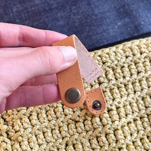 May include: Close-up of a light brown leather strap with a brass buckle attached to a light green crocheted bag.