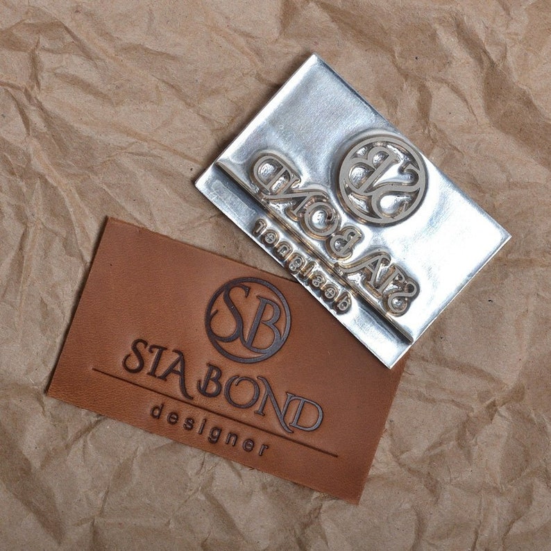 May include: A close-up of a metal stamp with the text "Sia Bond" and a circle with the letters "SB" inside. The stamp is on top of a piece of brown leather with the same design stamped into it.