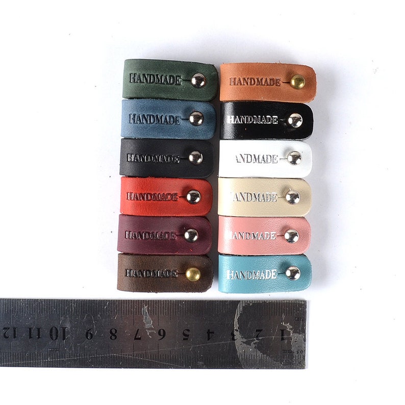 Op de afbeelding: Een set van 12 leren kabelbinders in verschillende kleuren, elk met een zilveren of gouden metalen klinknagel en het woord "HANDMADE" gegraveerd op het leer.