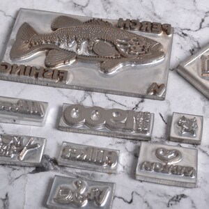 May include: A collection of metal stamps with various designs, including a fish, the word "fishing", the word "perch", the number "1900", the word "biker", the word "love", and a heart. The stamps are on a white marble surface.