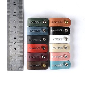 Op de afbeelding: Acht leren kabelbinders in verschillende kleuren, elk met een zilveren of gouden metalen knoop en het woord "HANDMADE" gegraveerd op de voorkant.