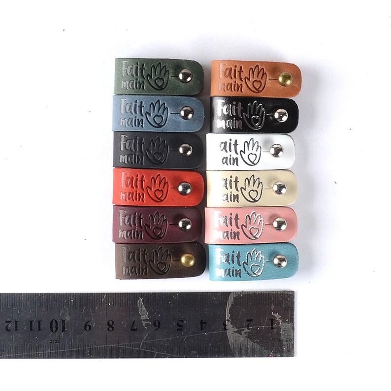 May include: A set of 10 leather tags with the words "Fait Main" and a heart-shaped hand design. The tags are in various colors, including green, blue, black, white, red, burgundy, and teal.