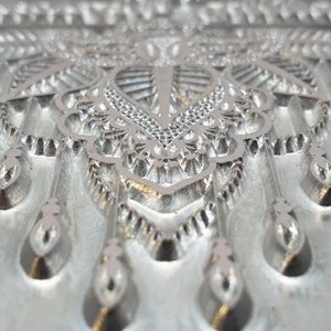 May include: A close-up of a silver metal mold with a detailed, intricate design. The mold features a repeating pattern of teardrop shapes and geometric designs.