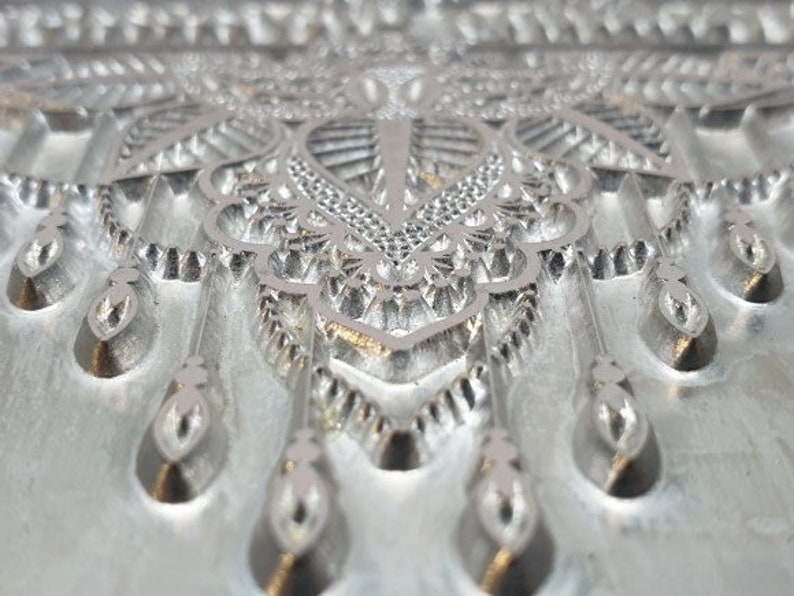 May include: A close-up of a silver metal mold with a detailed, intricate design. The mold features a repeating pattern of teardrop shapes and geometric designs.