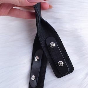 Puede incluir: Primer plano de dos correas de cuero negro con hebillas de metal plateado. Las correas est&aacute;n cosidas juntas y tienen una superficie texturizada.