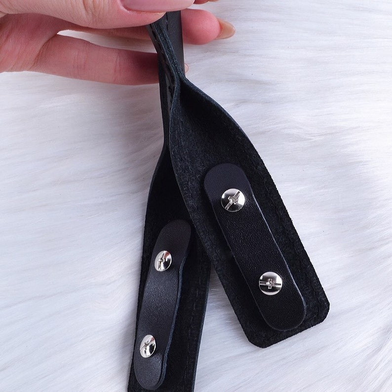 Puede incluir: Primer plano de dos correas de cuero negro con hebillas de metal plateado. Las correas est&aacute;n cosidas juntas y tienen una superficie texturizada.