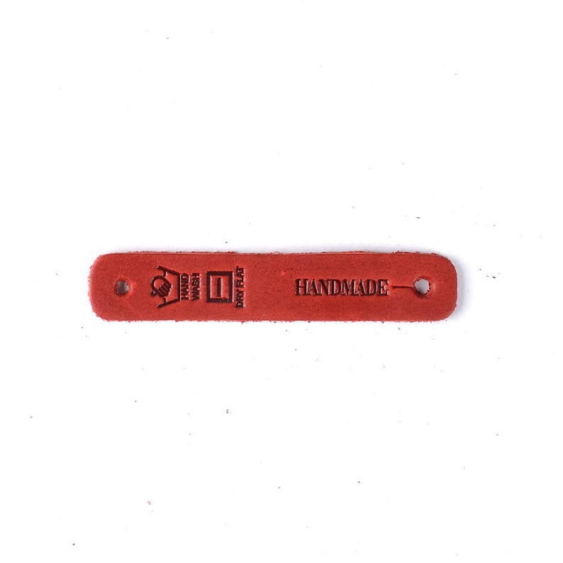 Op de afbeelding: Rood leren label met "HANDMADE" en wasinstructies: "HAND WASH", "DRY FLAT".