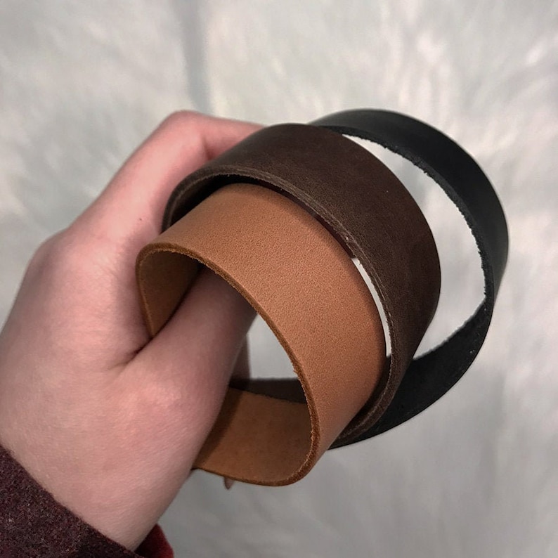 May include: Three leather straps in different colors: brown, tan, and black. The straps are rolled up and held in a hand.