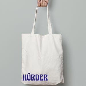 May include: A white canvas tote bag with long handles is held up by a hand. The bag features the word "HÜRDER" printed in blue, centered at the bottom. The background is a solid light gray.