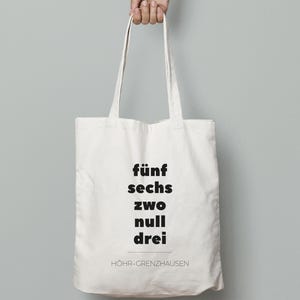 May include: A white canvas tote bag held by a hand. The bag features black text: "fünf sechs zwei null drei" and "HÖHR-GRENZHAUSEN" below. The bag has long handles and is against a light gray background.