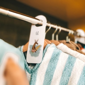 May include: A clothing rack with various items, including a blue and white striped garment. A tag with the number 68 and a cartoon deer surfing is attached to a wooden hanger. The image has a soft focus.