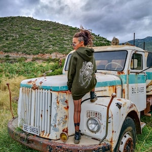 May include: A woman wearing a green hooded sweatshirt with a spider and skull graphic stands on the bonnet of a white, rusty truck. The truck has a number plate that reads "BE-9468".