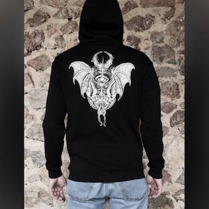 Puede incluir: Sudadera con capucha negra con un estampado blanco de un murciélago con un ojo y una luna creciente en la espalda.