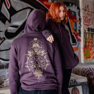 Puede incluir: Dos personas con sudaderas con capucha moradas con un diseño geométrico dorado en la espalda. La persona de delante lleva pantalones vaqueros negros y la persona de atrás lleva pantalones negros.