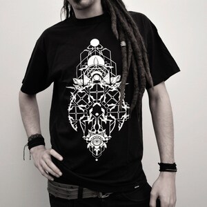 Op de afbeelding: Zwart T-shirt met een wit geometrisch ontwerp met een gestileerde boom en een cirkel met een kruis erin. Het ontwerp is gecentreerd op het shirt.