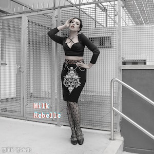 Puede incluir: Una mujer con un top negro de manga larga y cuello halter, una falda negra con estampado gráfico y medias de rejilla negras. La falda tiene un cinturón de cadena plateada con un dije de calavera. La mujer está de pie frente a una puerta de metal.