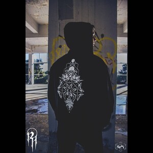 Op de afbeelding: Een zwart hoodie met een wit geometrisch ontwerp op de rug. Het ontwerp bevat een gestileerd oog, een schedel en andere abstracte vormen.