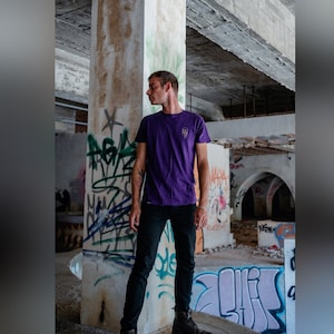 Op de afbeelding: Een persoon draagt een paars T-shirt met een klein grafisch ontwerp op de voorkant, een zwarte jeans en zwarte schoenen. De persoon staat in een gebouw met graffiti op de muren.
