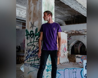May include: A person wearing a purple t-shirt with a small graphic design on the front, black jeans, and black shoes. The person is standing in a building with graffiti on the walls.
