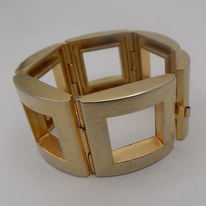 Puede incluir: Una pulsera dorada con un diseño geométrico. La pulsera está compuesta por eslabones rectangulares con centros cuadrados abiertos, conectados por bisagras. El metal tiene un acabado cepillado y la pulsera se muestra sobre un fondo blanco.