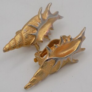Könnte beinhalten: Ein Paar goldfarbene Clip-Ohrringe in Muschelform. Die Ohrringe haben eine strukturierte Oberfläche mit silbernen Akzenten. Das Design erinnert an eine Muschel, passend für Schmuck mit Strand-Thema.