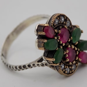 Könnte beinhalten: Ein silberner Ring mit einem Cluster aus roten und grünen Edelsteinen. Der Ring hat eine strukturierte Band und eine dekorative Fassung.