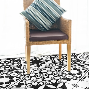 Puede incluir: Azulejos de suelo con estampado en blanco y negro con una silla de mimbre con un cojín a rayas en la parte superior. La silla está hecha de mimbre natural y tiene un cojín marrón. Los azulejos del suelo están dispuestos en un patrón de patchwork.