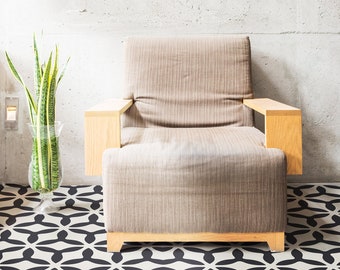 Puede incluir: Un sillón beige con brazos de madera se encuentra sobre un suelo con estampado geométrico en blanco y negro. Una planta en maceta está en el fondo.