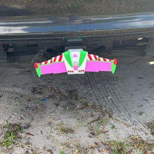 May include: A colorful, novelty trailer hitch cover shaped like a cartoon spaceship. The cover features a pink body with green and white accents, and red wing tips. It is attached to the hitch of a black vehicle, resting above a sandy surface.