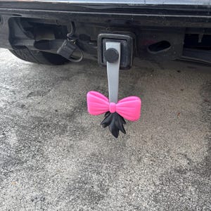 Puede incluir: Una cubierta de enganche de remolque en forma de lazo rosa con un acento negro, sujeta a un vehículo negro. El lazo es de color rosa brillante y tiene un diseño detallado. La cubierta es un accesorio divertido y decorativo.