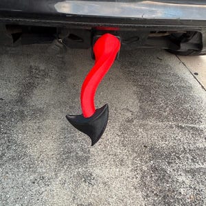 May include: A red and black devil tail hitch cover attached to a black vehicle. The tail is a curved, twisted design with a textured black arrow-shaped tip. The hitch cover adds a unique, decorative element to the vehicle's exterior.