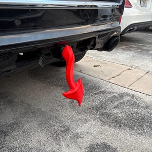 Puede incluir: Una cubierta de enganche de remolque con forma de cola de diablo roja brillante, fijada en la parte trasera de un SUV negro. La cubierta del enganche tiene forma de cola de diablo, añadiendo un detalle único y llamativo al exterior del vehículo.