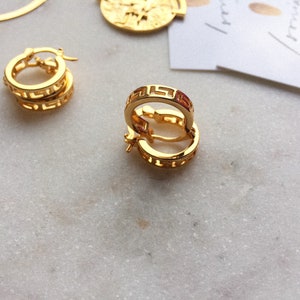 May include: A pair of gold hoop earrings with a Greek key design. The earrings are on a white marble surface.