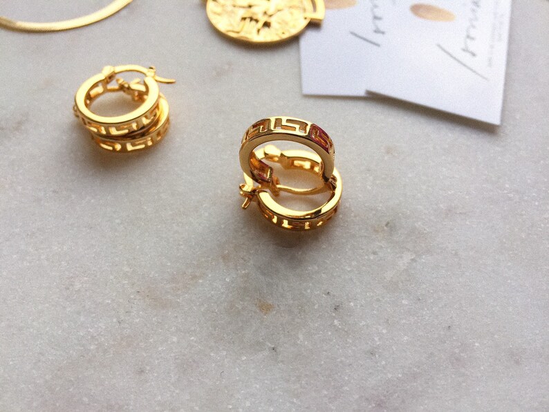 May include: A pair of gold hoop earrings with a Greek key design. The earrings are on a white marble surface.