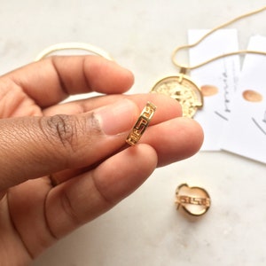 May include: Gold ring with a Greek key design. The ring is being held by a hand.